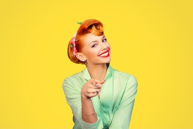 portrait of a beautiful woman pinup retro style pointing at you smiling laughing isolated yellow background wall. body language, gestures, psychology.