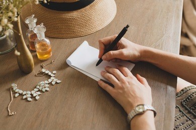 young stylish beautiful woman sitting at table in resort hotel room, writing a letter, holding pen, straw hat, vintage style, hands close-up, details, accessories, travel diary