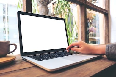 mockup image of hand using laptop with blank white screen on vintage wooden table in cafe
