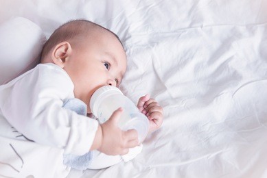 little asian child lying on white bed and drinking milk from bottle, holding bottle by himself.