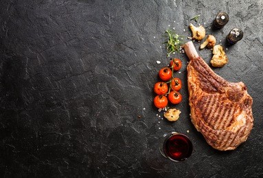 succulent grilled tomahawk beef steak on the bone with red wine, seasonings, fresh rosemary and grilled vegetables on a black background, top view