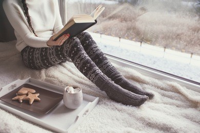 young woman with cup of coffee and gingerbread is reading a book sitting home relaxing by the window. snow winter home chill out concept