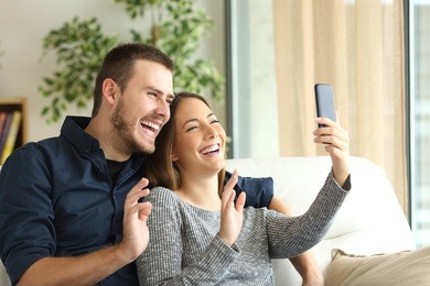 happy casual couple greeting in a phone video call on line sitting on a sofa in the living room at home with a window in the background