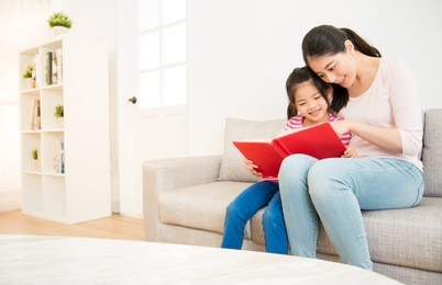 happy asian chinese mother and little daughter girl reading a book in the morning together in the living room at home. family activity concept.