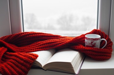 cozy winter still life: mug of hot tea and opened book with warm plaid on modern windowsill against snow landscape from outside. selective focus.