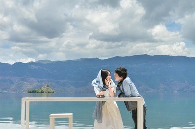 happy young chinese couple leans on white long table looking at each other, by the bank of lugu lake, at daluoshui village, lijiang, china