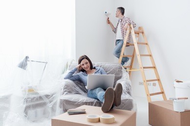 happy couple doing home renovations, the man is painting the room and the woman is relaxing on the armchair and connecting with a laptop