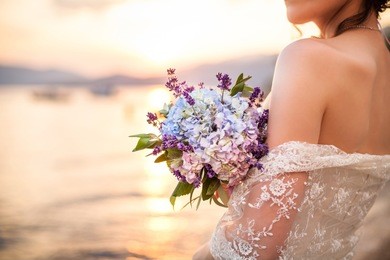 the bride is holding wedding bouquet from lavender and hydrangea and standing on background of the sea and sunset after wedding ceremony.