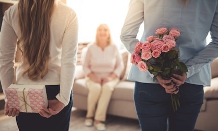 teenage girl and her mom are hiding flowers and a gift box for their beautiful granny behind backs while grandma is sitting on couch at home