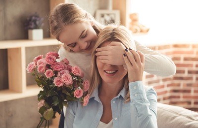 cute teenage girl is covering her mom's eyes while giving flowers and a gift box to her