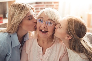 three generations of women. beautiful woman and teenage girl are kissing their granny while sitting on couch at home