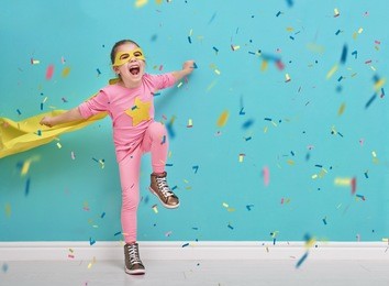 little child plays superhero. kid on the background of bright blue wall. girl is throwing confetti and jumping. yellow, pink and  turquoise colors.
