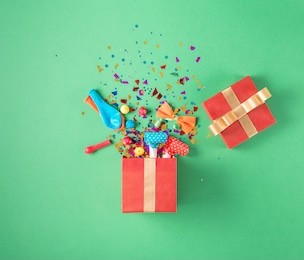 red gift box with various party confetti, balloons, streamers, noisemakers and decoration on a green background. colorful celebration background. flat lay.