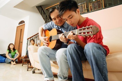 father teaching son to play guitar, mother and daughter in the background
