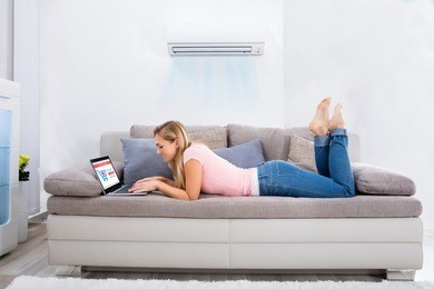 young happy woman lying under air conditioner on couch using laptop at home