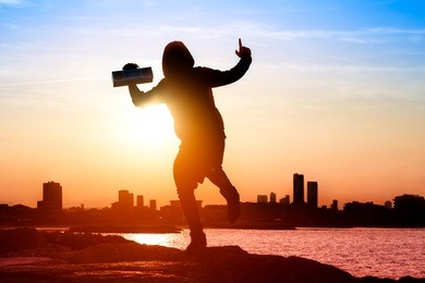 silhouette of happy man listening music radio tape player on cityscape background at sunset - hooded guy figure dancing on the beach with joyful winner attitude backlight image and sun halo filter