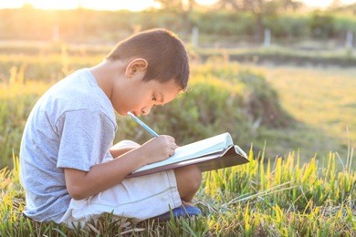 little asian boy writing book with pencil in the park on sunset