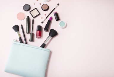 a blue cosmetics bag with makeup products spilling out on to a pastel pink background, with empty space at side