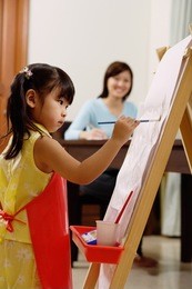 young girl painting on easel, mother in the background