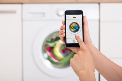 close-up of woman hand using mobile phone app to operate smart washing machine in kitchen