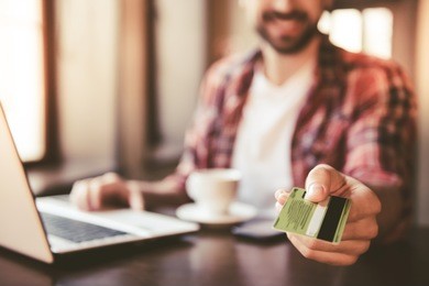 handsome man is giving a credit card and smiling while working with a laptop in cafe