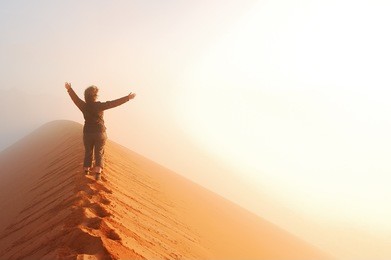 person standing on top of dune in desert and looking at rising sun in mist with hands up, travel in africa, namibia
