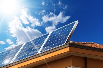 close-up of a house roof with a solar panels on top, on a blue sky with clouds and sun rays