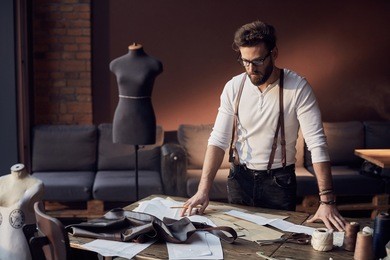cute tailor male with beard and glasses in white shirt with brown leather suspenders working near wooden table with threads, apron and scissors in amazing atelier with antique furniture 