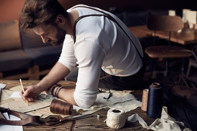 handsome tailor male with beard in white shirt with brown leather suspenders working near wooden table with threads in amazing atelier with antique furniture and mannequin on background