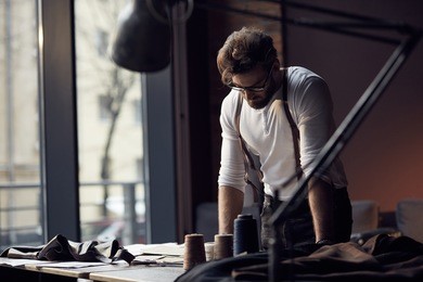 young serious tailor with beard and glasses in white shirt with leather suspenders looking on drawing near wooden table with threads in amazing atelier with antique furniture and mannequin 