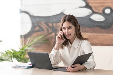 good business talk. cheerful young asian girl beautiful asian woman talking on mobile phone and using laptop with smile while sitting at her working place