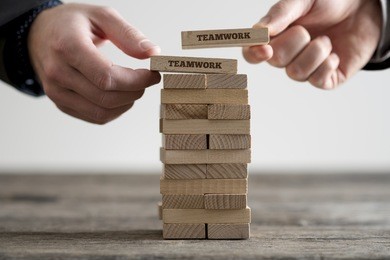 two hands putting dominoes onto stack of wooden bricks with teamwork business signs on rustic table surface.