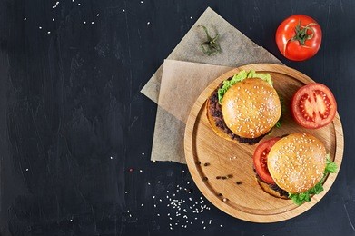 craft beef burgers on round wooden cutting board with vegetables. flat lay on black textured background.
