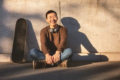 asian handsome young man sitting with back against wall,shadows and a skateboard beside,headphone around neck