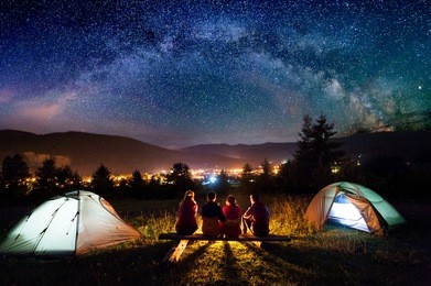 friends hikers sitting on a bench made of logs and watching fire together beside camp and tents in the night. on the background beautiful starry sky, mountains and luminous town. rear view