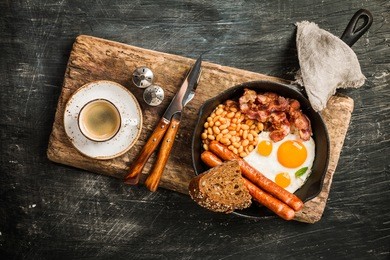 english breakfast - fried egg, beans, tomatoes, sausages, bacon and bread in frying pan on aged board over black background. top view