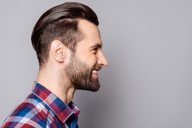 a side view portrait of young handsome smiling man with stylish haircut standing against gray background.