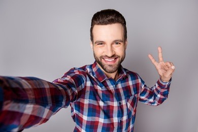 young funny smiling man isolated on gray background taking a selfie and showing v-sing in camera.