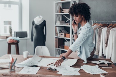 in touch with suppliers. beautiful young african woman talking on smart phone while sitting on desk in her studio with clothes hanging in the background 