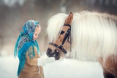 russian style. beautiful little girl in blue pavloposad shawl is feeding pony in winter in russia. image with selective focus and toning