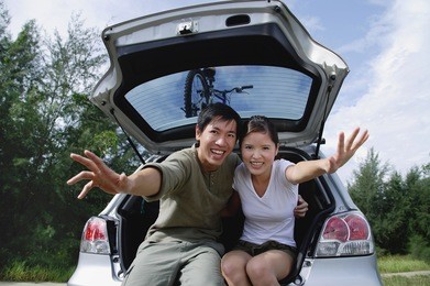 couple sitting in boot of car, reaching towards camera, smiling