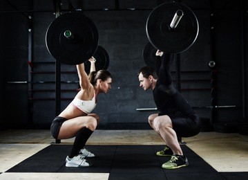 motivational side view image of young man and woman performing overhead squats with huge heavy barbells looking at each other