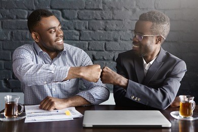 candid shot of happy successful dark-skinned businessmen wearing formal clothing fist-bumping while cheering and congratulating each other after making profitable deal and signing good contract