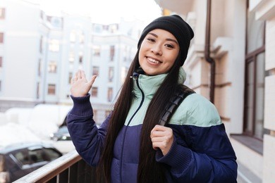 young smiling asian woman in warm clothes and with backpac which standing on the street, looking at waving at camera