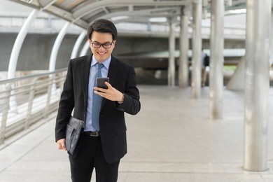 young asian handsome businessman and glasses using smartphone while walking outside business district before work travel with bag and smiling at business district 