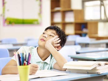 asian elementary schoolboy thinking while doing homework in classroom, looking up hand on cheek.