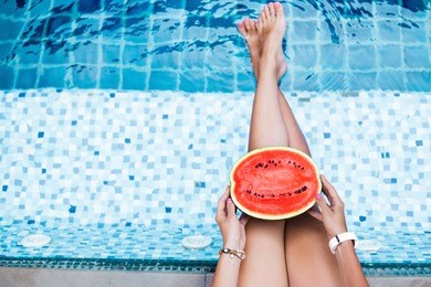 a girl holds half a red watermelon stretching long, tanned legs over a blue pool, relaxing on a tropical island in a hotel, eating healthy, fruit diet, summer style