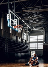 portrait of black basketball player sits on a floor under  a hoop in a basketball hall.