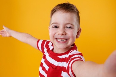 portrait of a satisfied cheerful little boy holding mobile phone and making selfie isolated over orange background