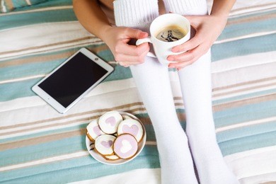 woman using tablet at cozy home atmosphere on the bed. young woman with cup of cocoa or coffee in hands enjoying free time with comfort. soft light and comfy lifestyle concept. top view point.
